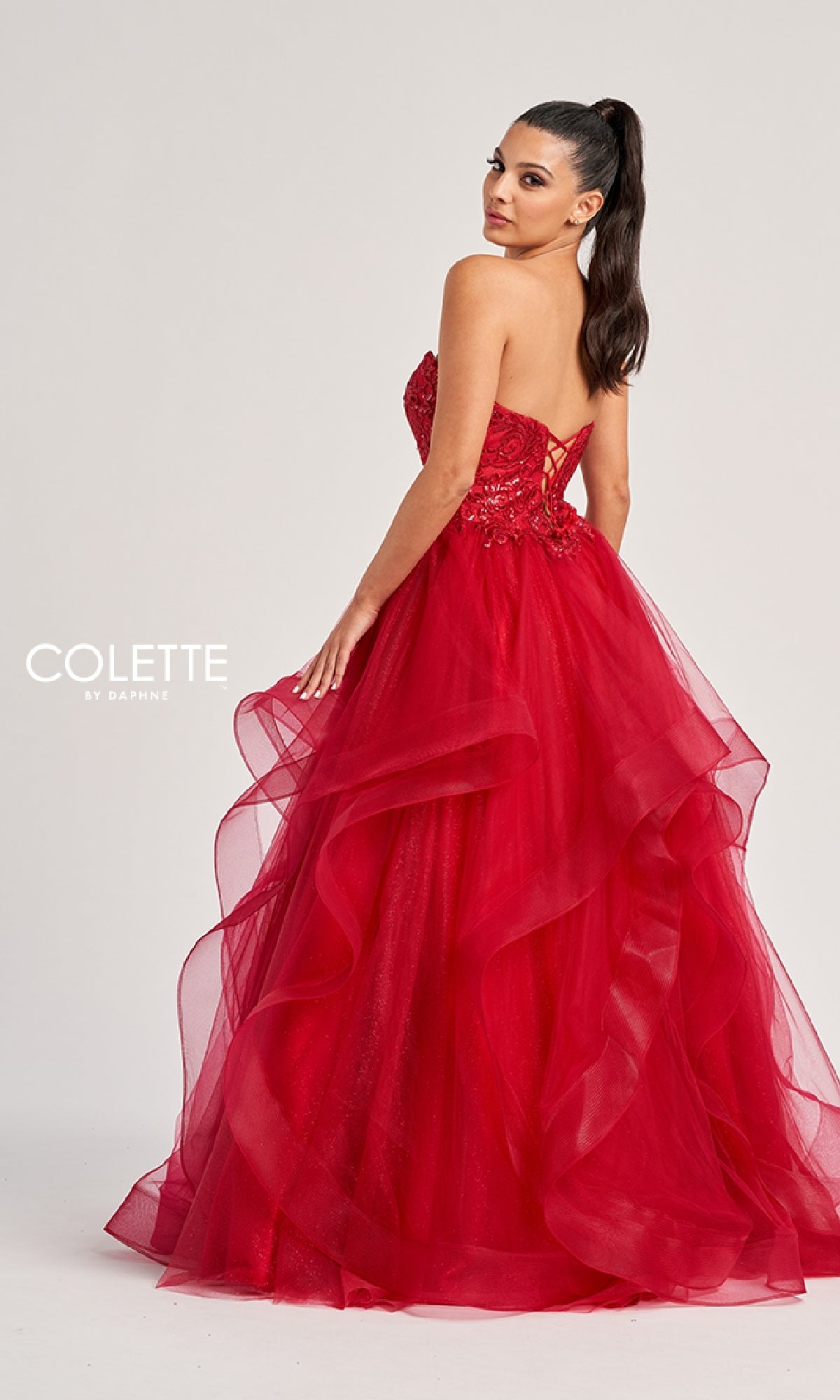 A woman poses with her head turned over her shoulder in the Colette CL8290 long prom dress, featuring a strapless bodice, bright red layered tulle skirt, and lace-up back.